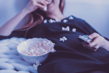 Mujer relajándose en casa viendo series en la televisión mientras come palomitas en el sofá.