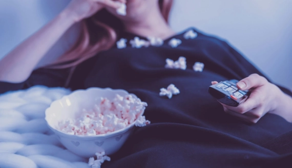 Mujer relajándose en casa viendo series en la televisión mientras come palomitas en el sofá.