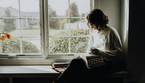 mujer adulta leyendo junto a la ventana con una taza de café, disfrutando la tranquilidad de vivir sola al independizarse