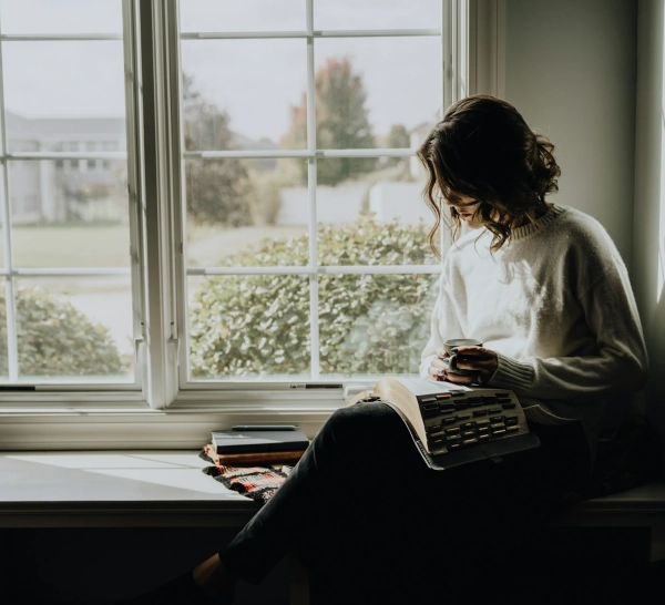 mujer adulta leyendo junto a la ventana con una taza de café, disfrutando la tranquilidad de vivir sola al independizarse